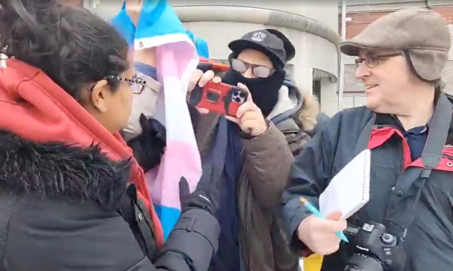 Guy with fogged up glasses pointing his camera at Caryma filming while he shoves someone holding a trans flag, with some older white guy holding a notebook next to them.